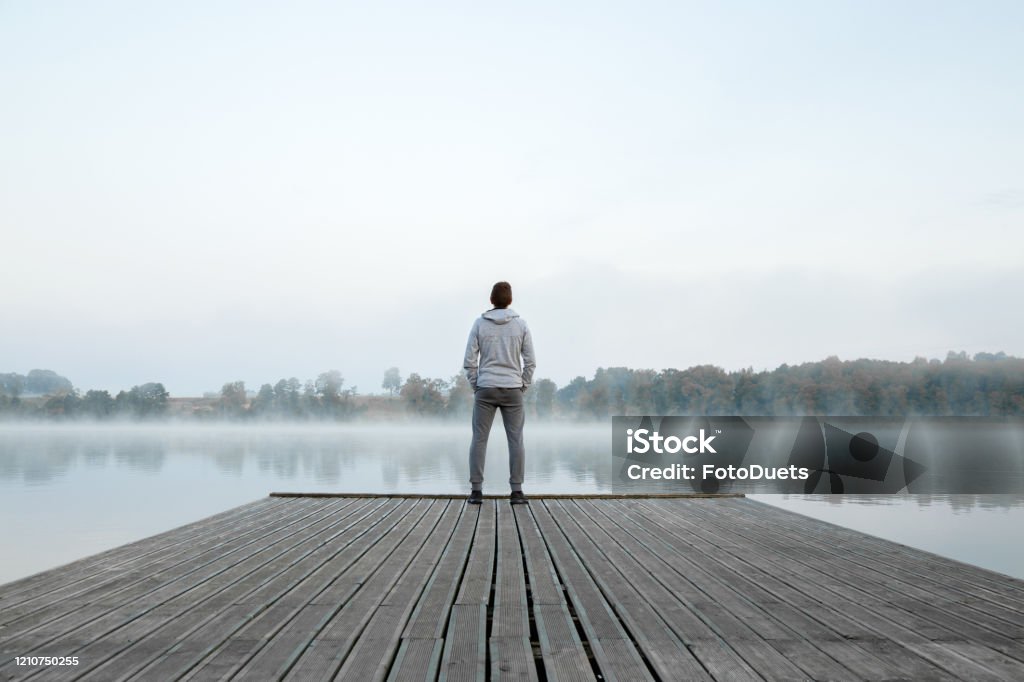 Young man standing alone on wooden footbridge and staring at lake. Thinking about life. Mist over water. Foggy air. Early chilly morning. Peaceful atmosphere in nature. Enjoying fresh air. Back view. Men Stock Photo