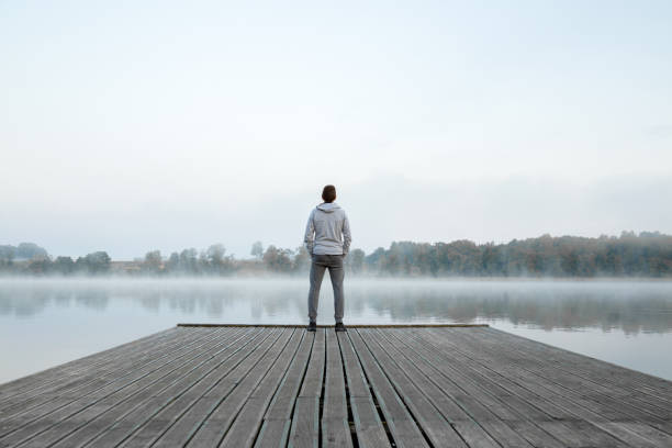 young man standing alone on wooden footbridge and staring at lake. thinking about life. mist over water. foggy air. early chilly morning. peaceful atmosphere in nature. enjoying fresh air. back view. - jetty imagens e fotografias de stock