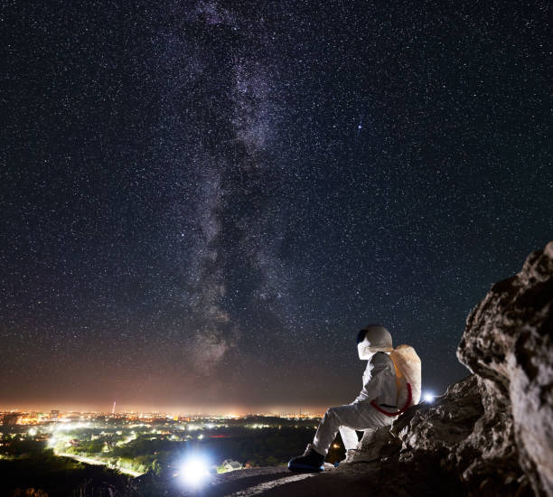 istock Spaceman sitting on rocky hill under magnificent night sky with stars. 1210750409