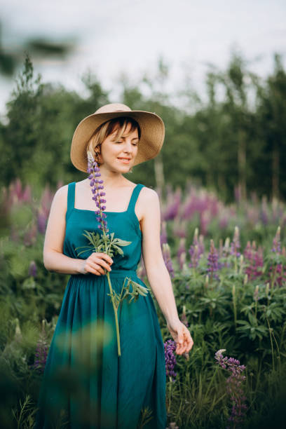 schöne romantische mädchen blondine in einem hut und kleid in einem feld von lila blumen von lupinen mit einer geheimnisvollen blume. - lupine single flower flower blue stock-fotos und bilder