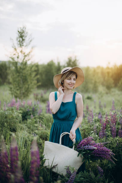 schöne romantische mädchen blondine in einem kleid und hut in einem feld mit einem strauß von lila blumen von lupinen in der morgendämmerung. - lupine single flower flower blue stock-fotos und bilder