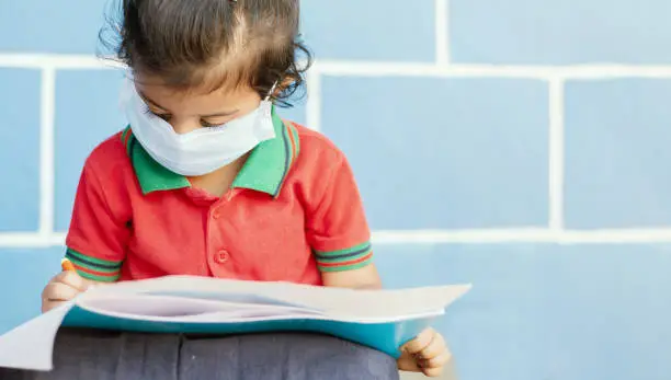 Covid 19 or Coronavirus and Air pollution pm2.5 concept - Little girl wearing medical mask and busy in writing at school - showing Wuhan covid-19 or sars cov 19 outbreak or epidemic of virus