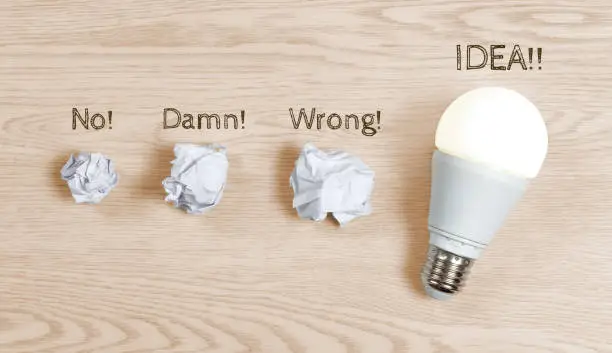 Three crumpled paper sheets and one switched-on lightbulb on a wooden background, to represent the coming to light of the right idea after a few attempts