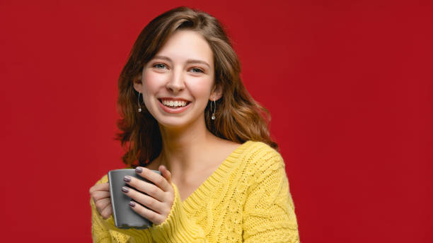 젊은 행복 한 백인 여자 따뜻한 음료의 회색 컵 노란색 스웨터를 착용. 빈 복사 공간이 있는 컨셉은 빨간색 배경이 있는 스튜디오에서 디자인에 대한 모형으로 - coffee women tea russian culture 뉴스 사진 이미지