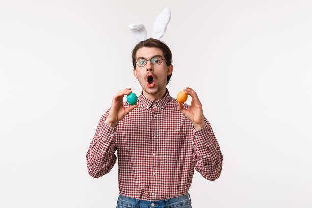 traditions, religious holidays, celebration concept. funny joyful young man with beard in glasses, wear cute rabbit ears and hold two painted eggs, playing game on easter day, white background - mask religious celebration horizontal easter imagens e fotografias de stock