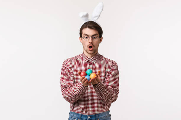 traditions, religious holidays, celebration concept. amused and excited caucasian young man in rabbit ears, glasses, open mouth amazed, holding painted eggs celebrating holy easter day - mask religious celebration horizontal easter imagens e fotografias de stock