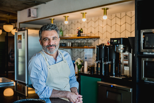 Bearded barista wearing apron and leaning on counter of corporate coffee bar in Buenos Aires office and smiling at camera.