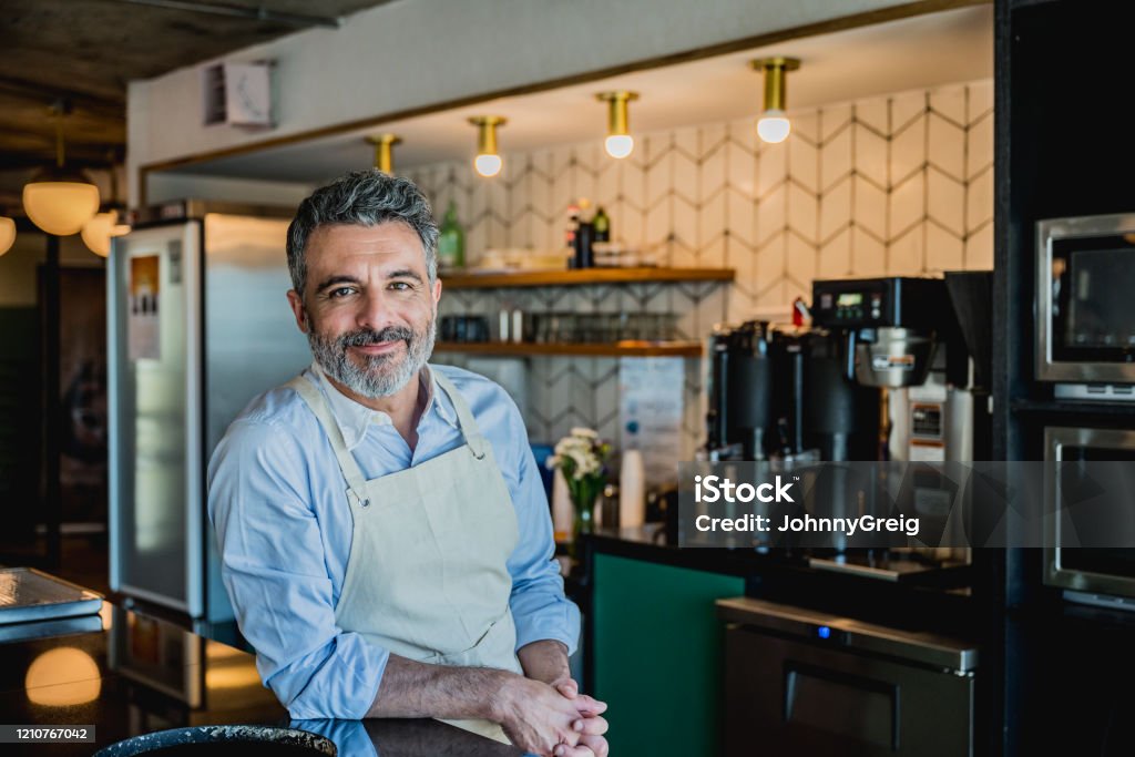 Smiling male barista ready to prepare drink in coffee bar Bearded barista wearing apron and leaning on counter of corporate coffee bar in Buenos Aires office and smiling at camera. Small Business Stock Photo