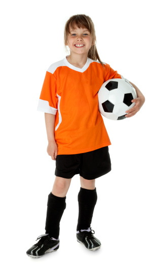 Little brunette girl with long hair in a ponytail wearing a large orange and white jersey and black shorts.  The girl is holding a soccer ball and standing in front of a white background.
