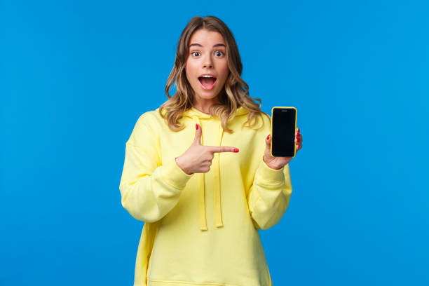 istock Impressed happy cheerful blond girl explain something as pointing mobile phone display, talking about new application or sharing her vacation photos, hold smartphone, look camera 1210773466