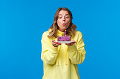 Celebration, party and lifestyle concept. Silly cute european blond woman in yellow hoodie, making wish holding fruit cake blowing out b-day candle, standing blue background at birthday party