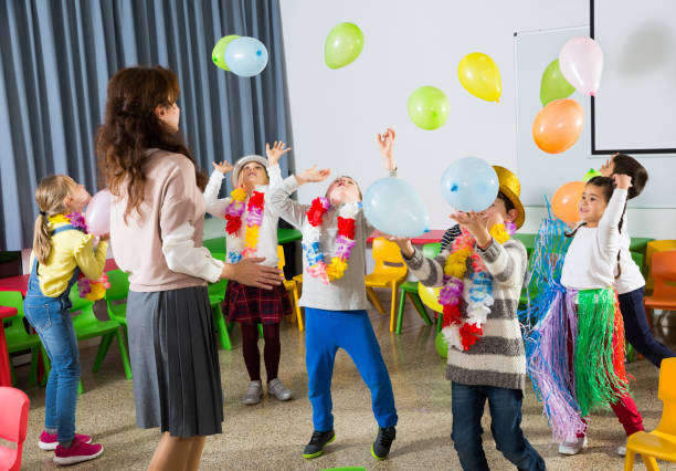 Happy kids and female teacher in funny hats and festive accessories playing together with colorful balloons in schoolroom