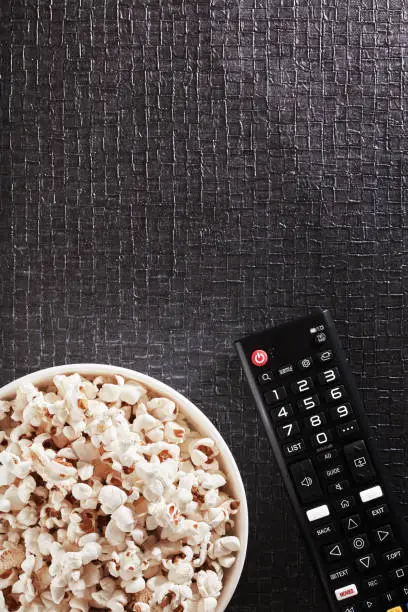 Photo of Bowl of popcorn with TV remote control on a black textured background