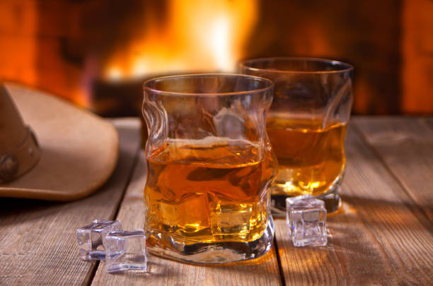 whiskey with ice on a wooden table with fireplace and cowboy hats on the background. stock photo