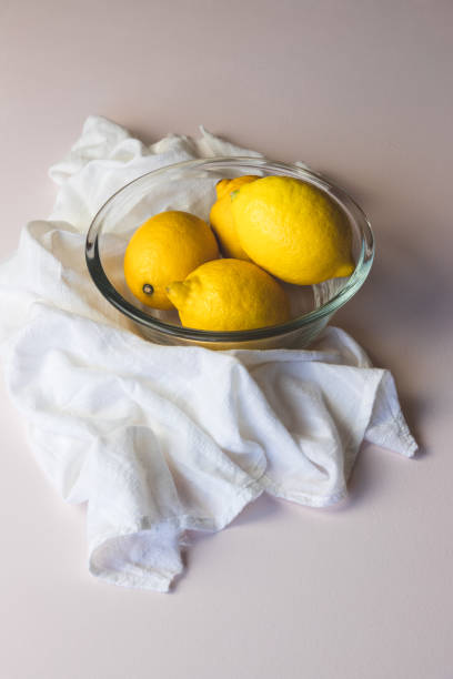 Lemons in a glass bowl, surrounded by a white cotton drape, on a light baby pink background