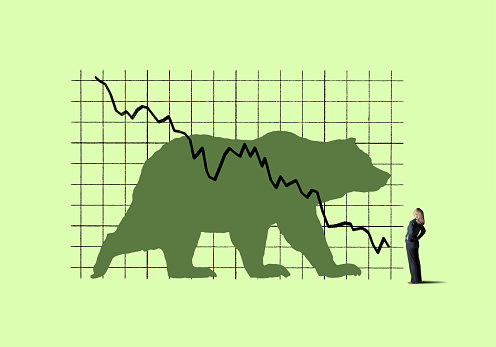 A businesswoman places her hand on her hips as she looks up at a large chart that indicates declining performance and has bear on it.