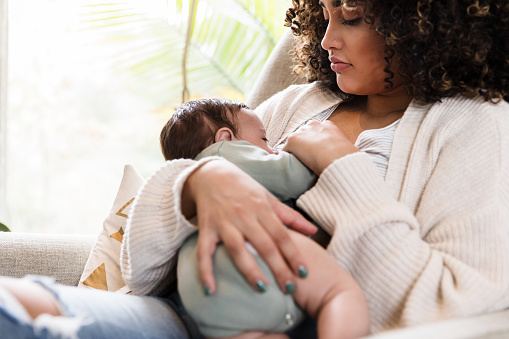 Sitting on a chair in the living room, the new mom watches as her baby boy breastfeeds.