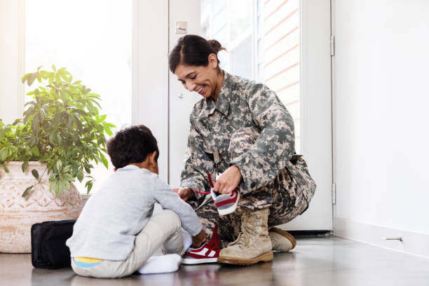 Mid adult female soldier kneels down to help her little boy put on his shoes before he leaves for a day at preschool.