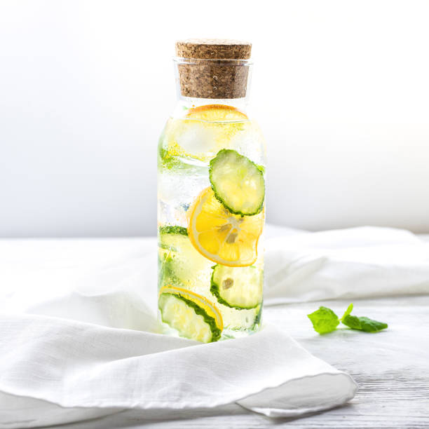 Detox Sassy water with lemon, cucumber and mint. A glass bottle of clean, cool and fresh drink stands on a white wooden table with a cotton natural napkin. Beautiful and enjoyable diet.