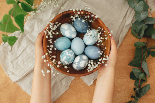 istock Hands holding wooden bowl with modern Easter eggs in spring flowers and eucalyptus on rustic table. Stylish pastel blue Easter eggs painted in natural dye from red cabbage. Happy Easter greetings 1210816245