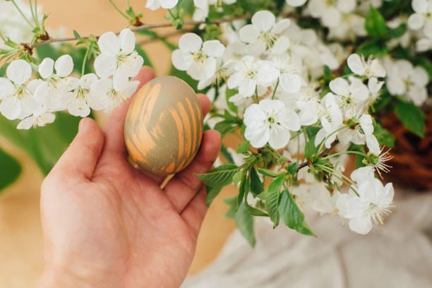 istock Happy Easter. Hand holding modern Easter egg on background of blooming spring flowers on rustic table with basket. Stylish pastel green Easter egg painted in natural dye.  Easter hunt 1210821538