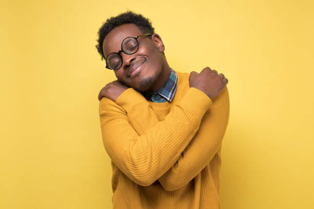 African american man in yellow clothes and glasses hugs himself Pleased african american man in yellow clothes and glasses hugs himself, has high self esteem. Studio shot on colored wall. one man only stock pictures, royalty-free photos & images