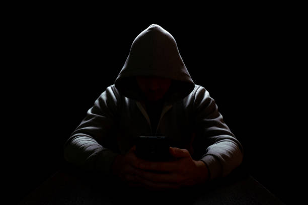 a man in a hood sits in complete darkness with a phone in his hands, selective focus
