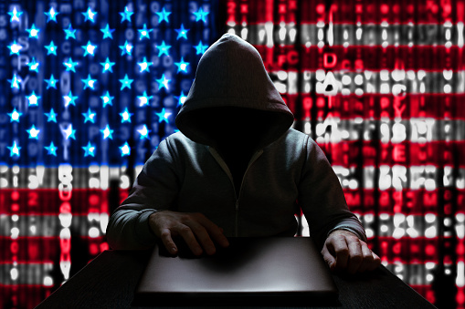 Cyberattack from the USA. An American hacker turns on a laptop, sitting at a table, on a background of binary code, the colors of the USA flag. DDoS attack