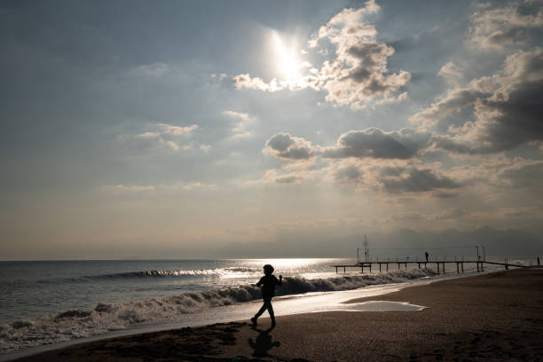 Full length photo of little boy playing with waves on beach sand during sunset. A pier is seen on the far end of horizontal frame. Sun and clouds are in view. Shot with a full frame mirrorless camera.