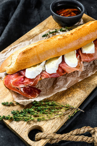 istock Baguette sandwich with prosciutto ham, Camembert cheese on a cutting Board. Black background, top view. 1210848116
