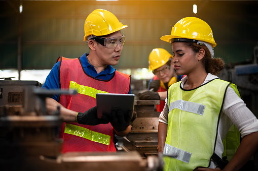 Young workers in industrial plants work in front of the machine. There is a consultation and conversation in the workplace. With a tablet And they wear uniforms, safety helmets and protective glasses. For work safety