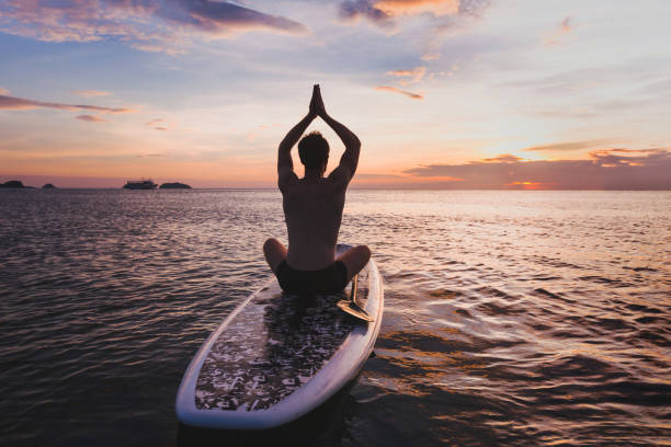 yoga su sup, silhouette dell'uomo seduto in posizione loto su stand up paddle board - sunset yoga young men caucasian foto e immagini stock