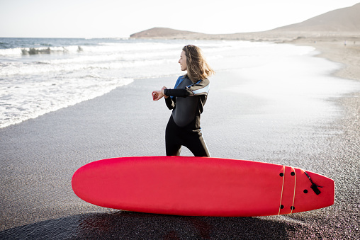 Young surfer in wetsuit standing with surf board on the wild beach on a sunset, looking on smart watch. Concept of using smart gadgets in sport