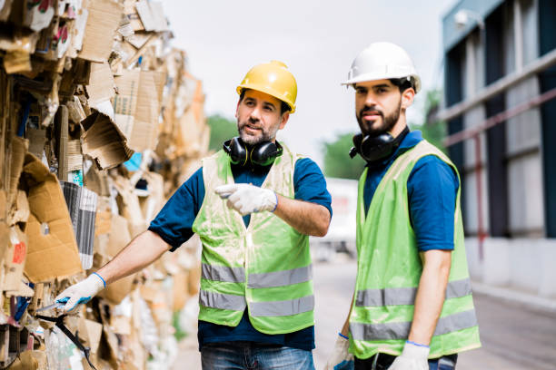 Male colleagues working at recycling factory Man discussing with coworker over stack of cardboard. Male workers are working at recycling factory. They are wearing protective workwear. recycling center stock pictures, royalty-free photos & images