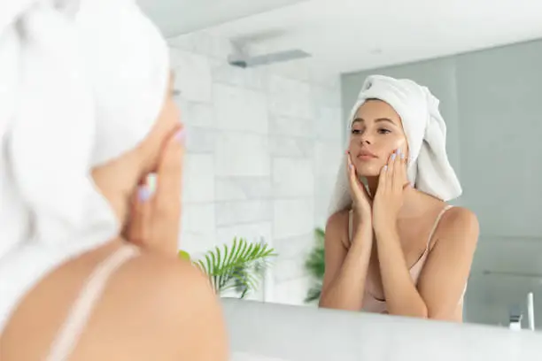 Young beautiful woman using skin face cream moisturizing lotion after taking bath. Pretty attractive girl wearing towel on head standing front of mirror in home bathroom. Daily hygiene and skincare