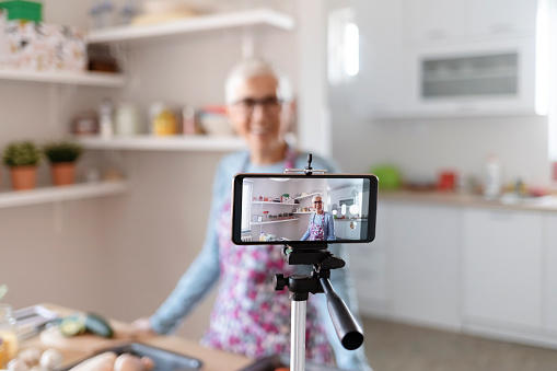 Senior Female Vlogger Making Social Media Video About Cooking for the Internet. Culinary Vlog. Baking Blog. Caucasian Mature Woman Cooking and Show Baking for Social Media. Video Recording at Kitchen. Focus on Mobile Phone
