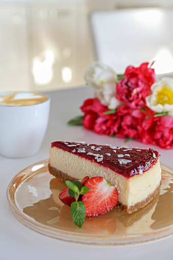 Delicious healthy vegetarian food, organic strawberry cheesecake slice, fresh red berries, green mint leaves, bouquet of different tulips and cup of cappuccino on white table. Close up, top front view