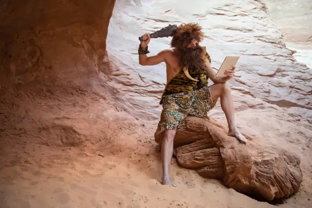 Stone Age luddite caveman scratching his head with a club while looking at his stone tablet outdoors in a weathered rock cave