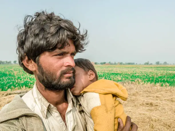 Photo of a young helpless father is carrying his cute and sleeping child on his shoulder and begging