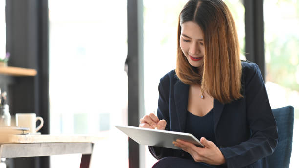 istock Executive woman holding a computer tablet and stylus pen in her hands while sitting at the modern working table with office glass wall as background. 1210948118