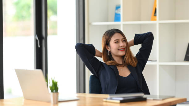 istock Woman relaxing after finished work while sitting at the modern working desk in front her computer laptop and books with modern white bookshelf and windows as background. 1210948122