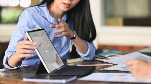 woman working as business development showing/talking about information graphs in computer tablet while sitting at the modern meeting table with orderly office as background. - discussion talking business explaining imagens e fotografias de stock