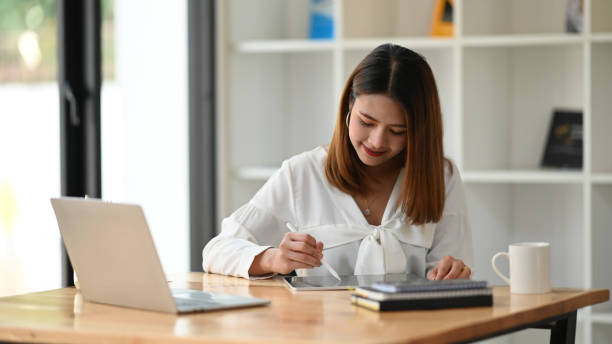 istock Woman holding stylus pen and using it to drawing on her computer tablet while sitting in front her laptop at the modern working table with office glass wall as background. 1210948144