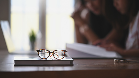 Glasses and book putting together on wooden table with two woman as blurred background. Library/Quiet place for reading book concept.