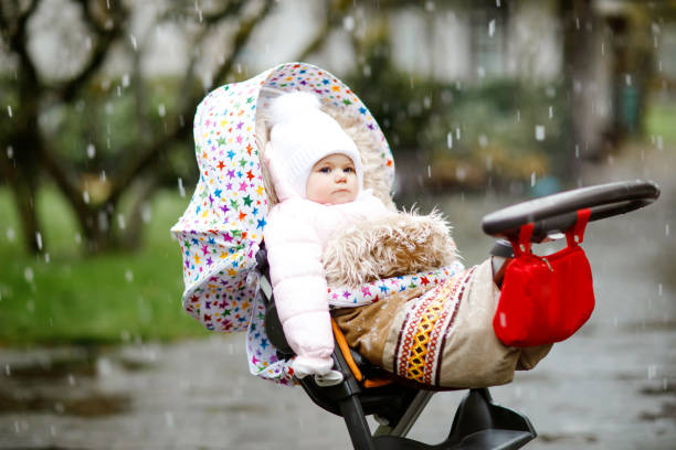 cute little beautiful baby girl sitting in the pram or stroller on cold day with sleet, rain and snow. happy smiling child in warm clothes, fashion stylish baby coat. winter or autumn day. - smiling little girls little boys autumn imagens e fotografias de stock