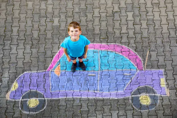 Photo of Adorable little kid boy playing with colorful chalks and painting big car picture on asphalt. Happy kid playing outside. Creative leisure for children outdoors in summer
