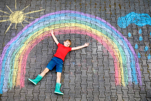 istock Happy kid boy in rubber boots with rainbow sun and clouds with rain drops painted with colorful chalks on ground or asphalt in summer. Creative leisure for children outdoors in summer 1210960487
