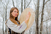 a woman in traditional clothing holds a shaman's drum, a white leather drum and wooden stick, during an outdoor music ceremony in the forest in the spring.