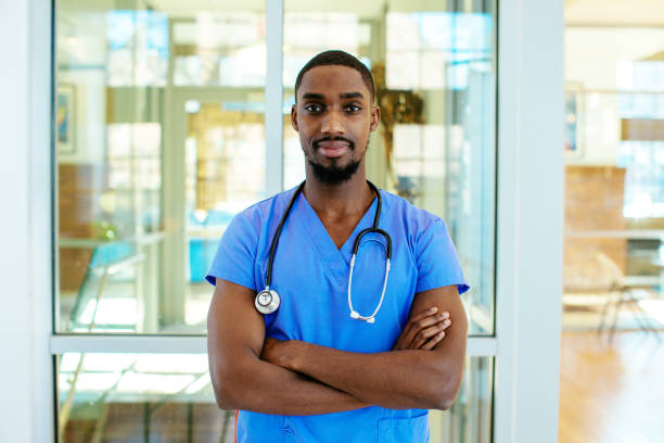 portrait of a serious young male doctor or nurse wearing blue scrubs uniform and stethoscope, with arms crossed in hospital - male nurse black nurse doctor imagens e fotografias de stock