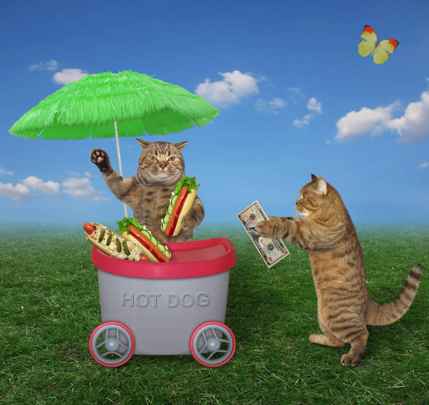 The beige cat is selling hot dogs in the grey mini movable kiosk under a green umbrella on the lawn. Butterflies are flying nearby.